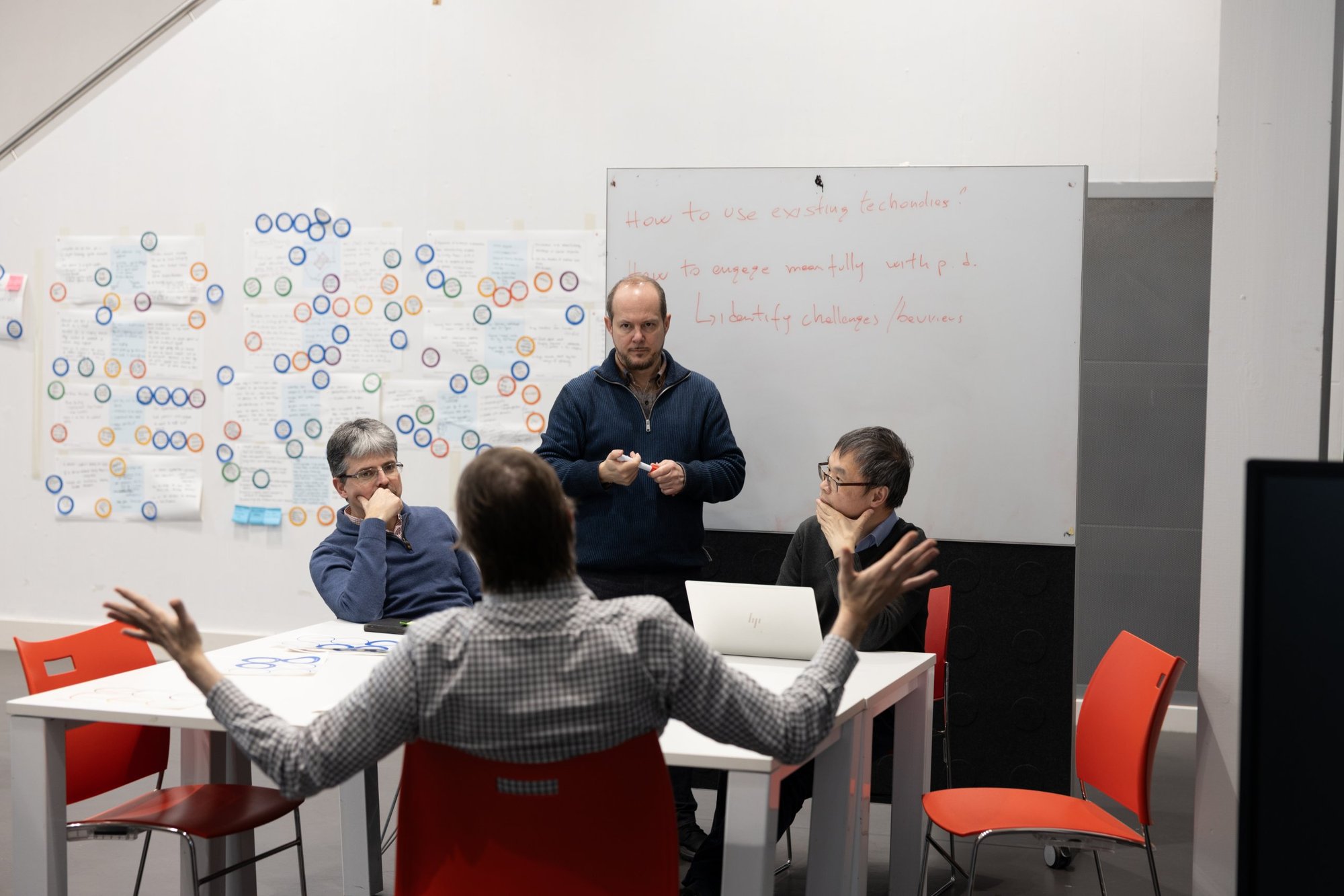 A small group discussing lived experience in front of a whiteboard and a wall covered in coloured sticker circles