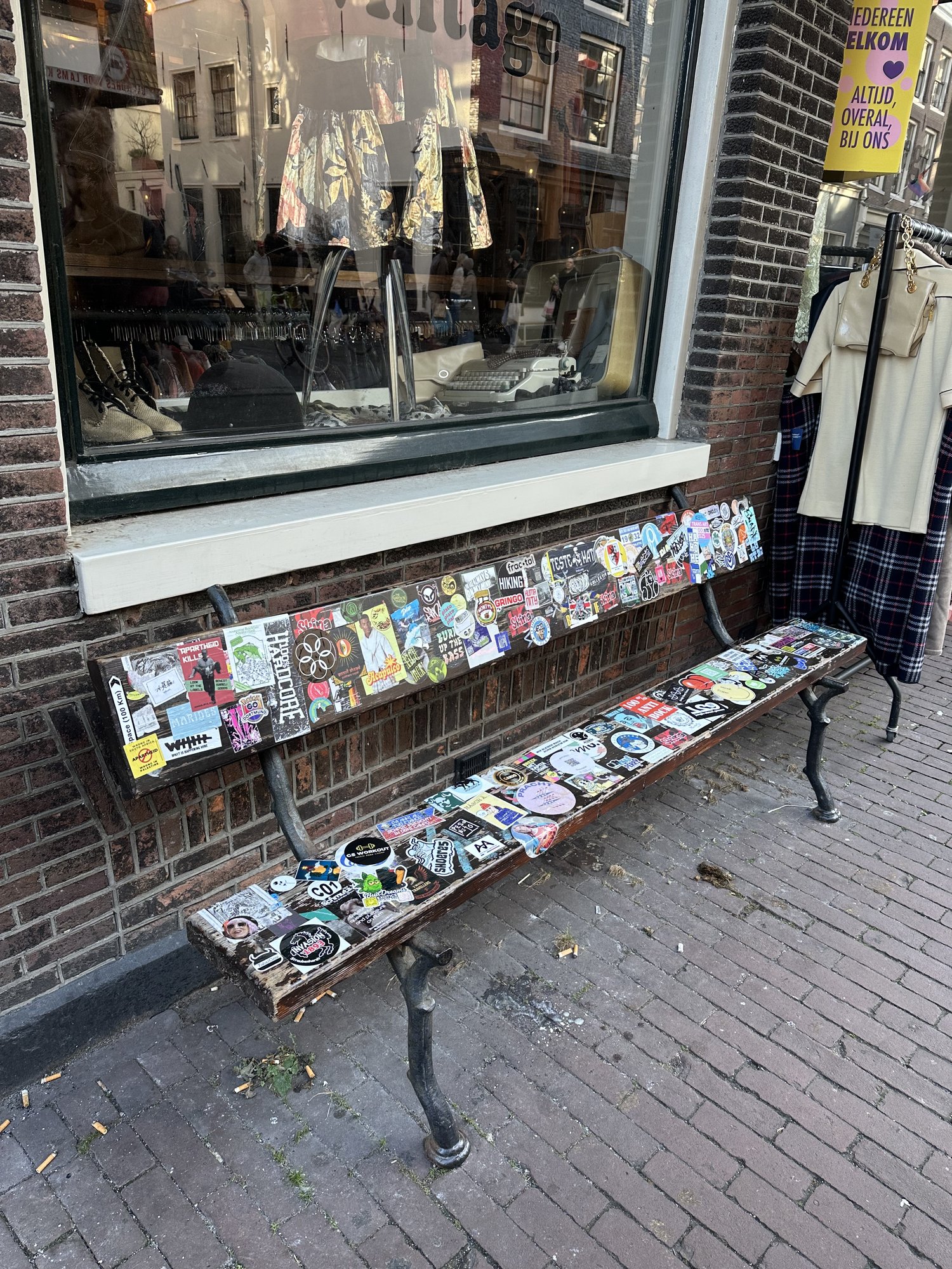 A sticker-covered bench outside a shop window on a brick street